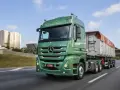 Zielony Mercedes Actros MP3 z naczepą na autostradzie, w tle budynki mieszkalne i most.