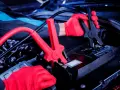 Mechanik w czerwonych rękawiczkach podłącza kable rozruchowe do akumulatora. Czy nowy akumulator trzeba ładować? Sprawdź instrukcję.