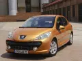 Pomarańczowy Peugeot 207 1.4 benzyna zaparkowany na brukowanym placu przed ceglanym budynkiem.