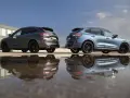 Dwa Fordy Kuga, jeden szary, drugi niebieski, stoją obok siebie na parkingu. Ich odbicia w kałuży tworzą surrealistyczny obraz. Oba modele to wersje hev.