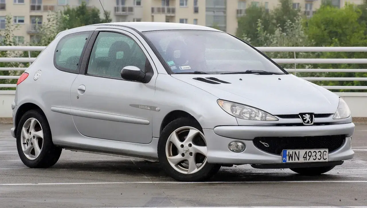 Srebrny Peugeot 206 Plus stoi na mokrym parkingu.
