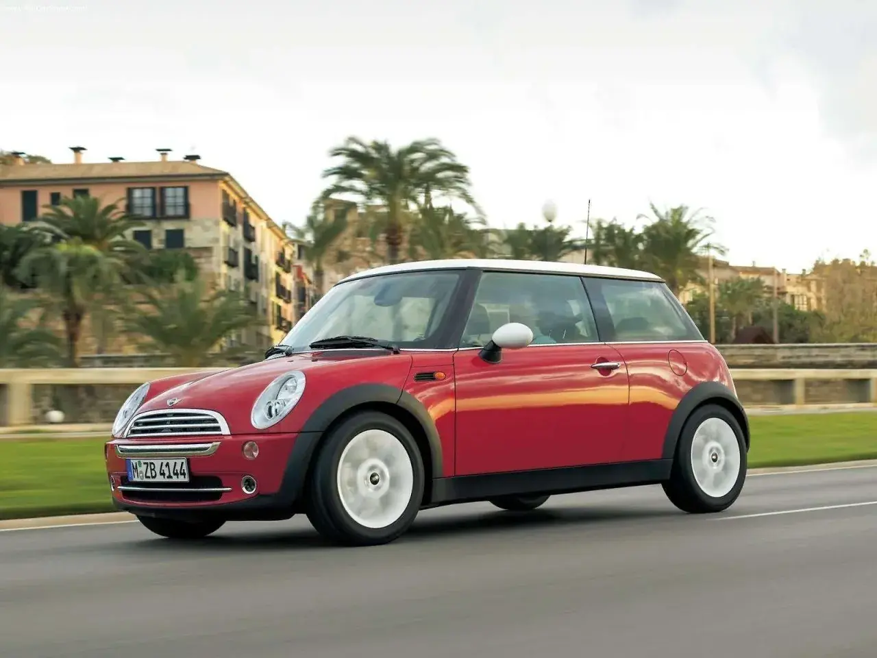 Czerwony mini r50 z białym dachem sunie po drodze, mijając palmy i budynki w słoneczny dzień.