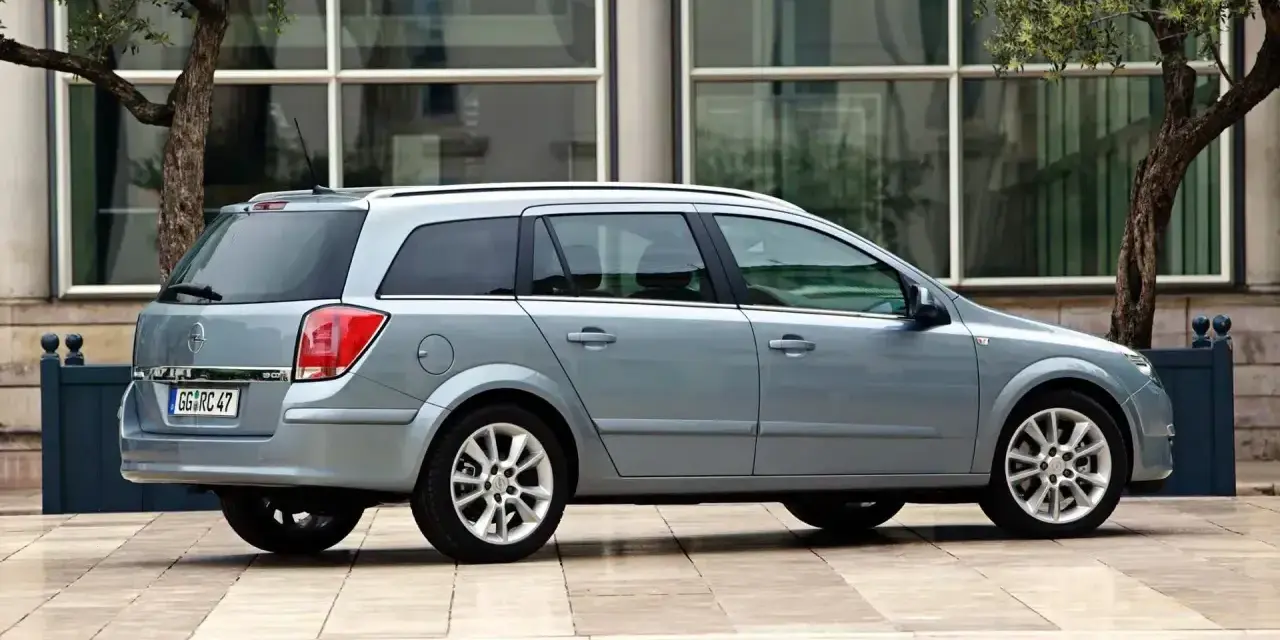 Srebrny Opel Astra H 1.6 benzyna kombi zaparkowany na tle nowoczesnego budynku z drzewami.