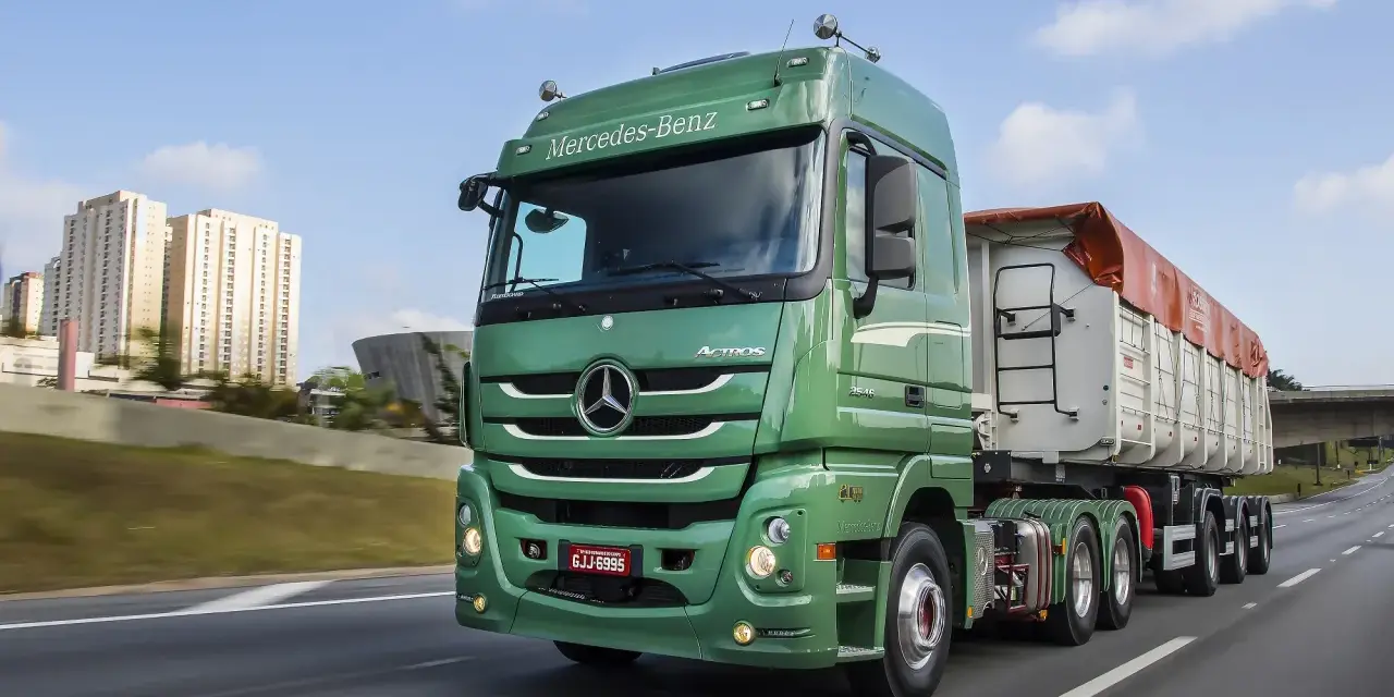 Zielony Mercedes Actros MP3 z naczepą na autostradzie, w tle budynki mieszkalne i most.