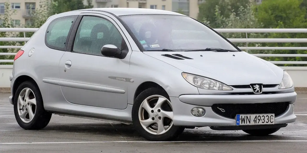 Srebrny Peugeot 206 Plus stoi na mokrym parkingu.