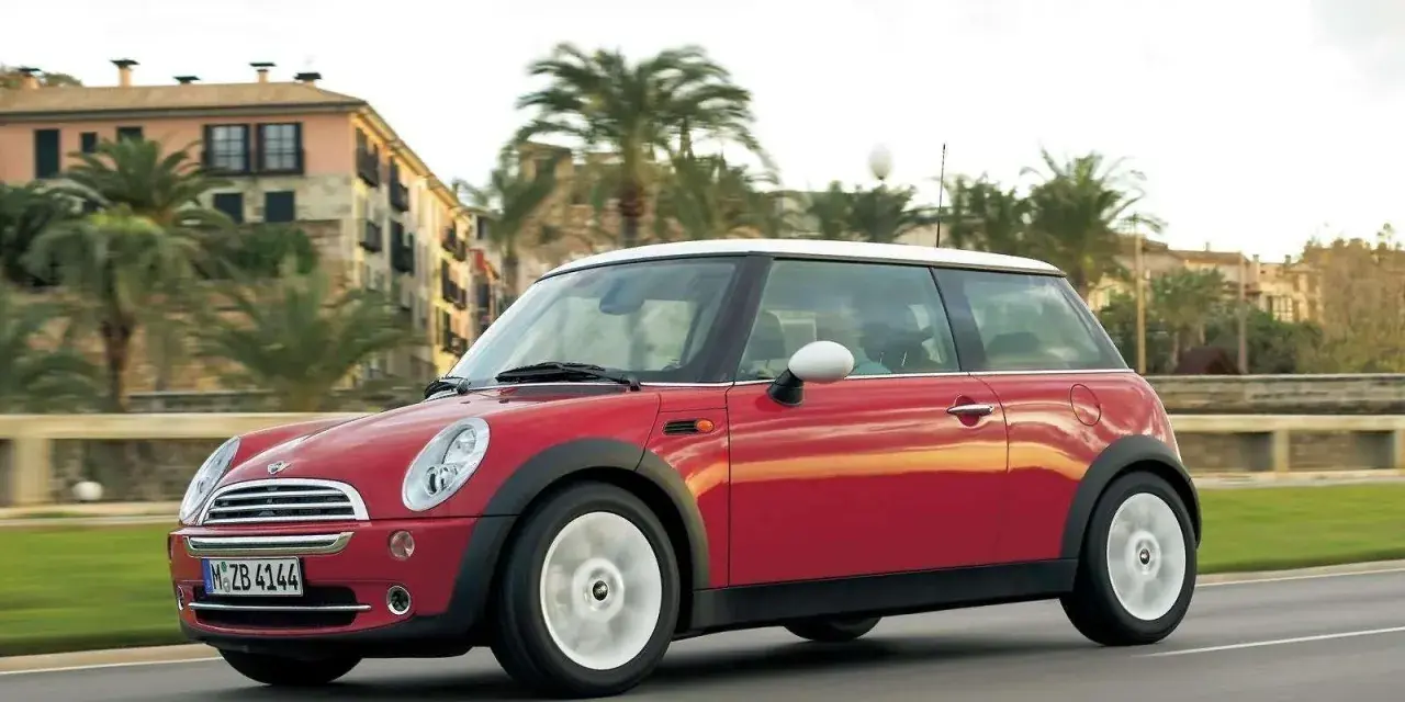 Czerwony mini r50 z białym dachem sunie po drodze, mijając palmy i budynki w słoneczny dzień.