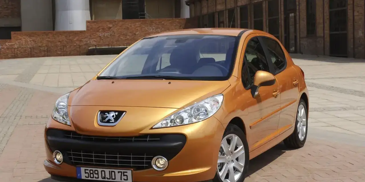 Pomarańczowy Peugeot 207 1.4 benzyna zaparkowany na brukowanym placu przed ceglanym budynkiem.