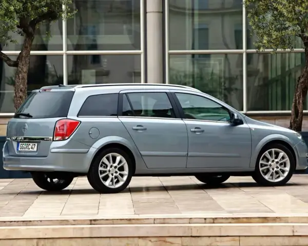 Srebrny Opel Astra H 1.6 benzyna kombi zaparkowany na tle nowoczesnego budynku z drzewami.