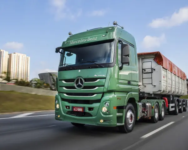 Zielony Mercedes Actros MP3 z naczepą na autostradzie, w tle budynki mieszkalne i most.