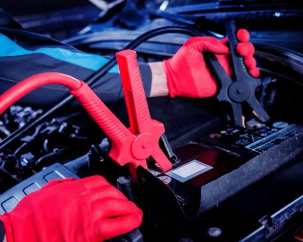 Mechanik w czerwonych rękawiczkach podłącza kable rozruchowe do akumulatora. Czy nowy akumulator trzeba ładować? Sprawdź instrukcję.