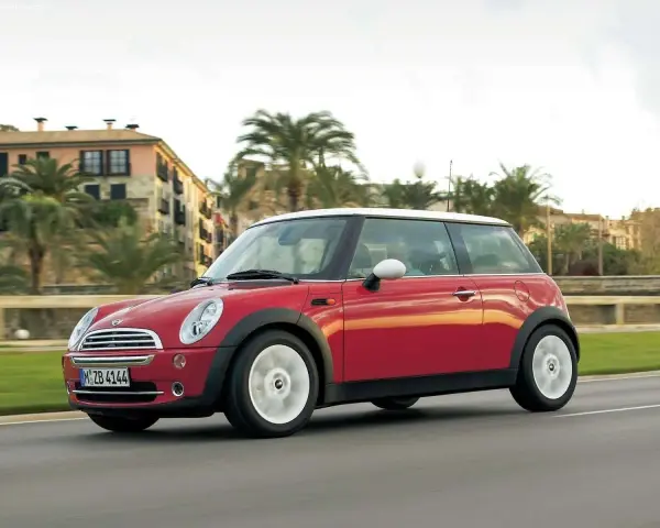 Czerwony mini r50 z białym dachem sunie po drodze, mijając palmy i budynki w słoneczny dzień.