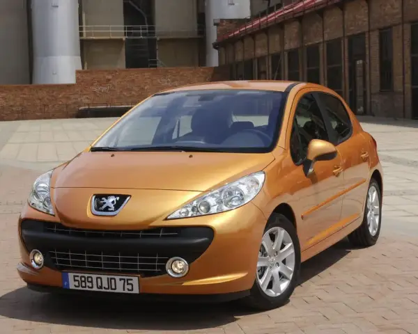 Pomarańczowy Peugeot 207 1.4 benzyna zaparkowany na brukowanym placu przed ceglanym budynkiem.
