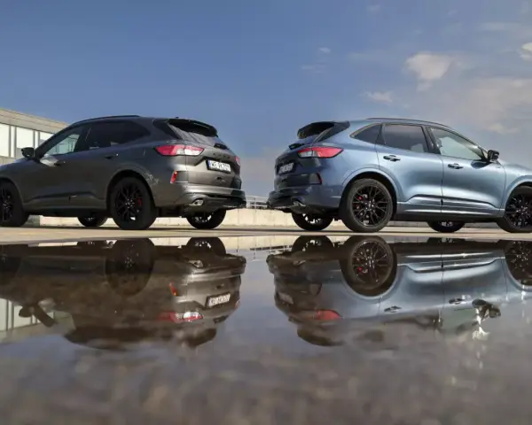 Dwa Fordy Kuga, jeden szary, drugi niebieski, stoją obok siebie na parkingu. Ich odbicia w kałuży tworzą surrealistyczny obraz. Oba modele to wersje hev.