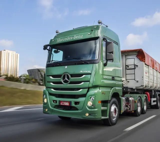 Zielony Mercedes Actros MP3 z naczepą na autostradzie, w tle budynki mieszkalne i most.