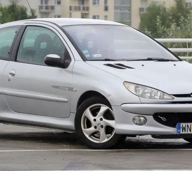 Srebrny Peugeot 206 Plus stoi na mokrym parkingu.