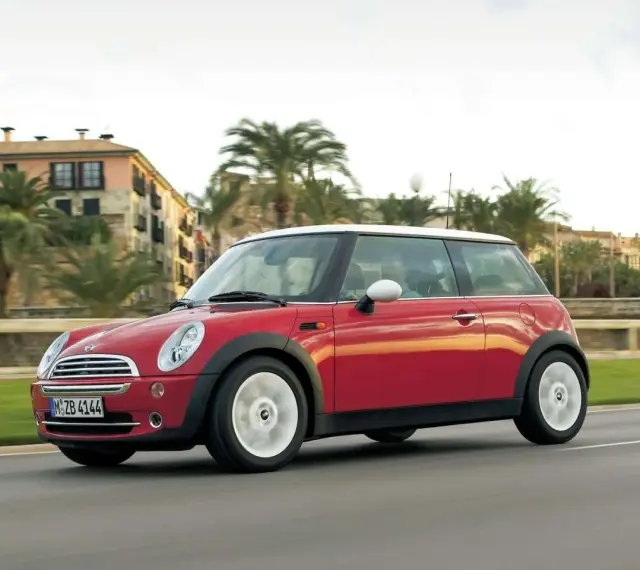 Czerwony mini r50 z białym dachem sunie po drodze, mijając palmy i budynki w słoneczny dzień.