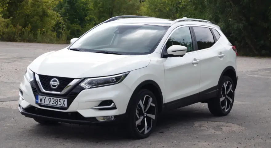 Biały Nissan Qashqai 2014 z polskimi tablicami rejestracyjnymi stoi na drodze, otoczony zielenią drzew.