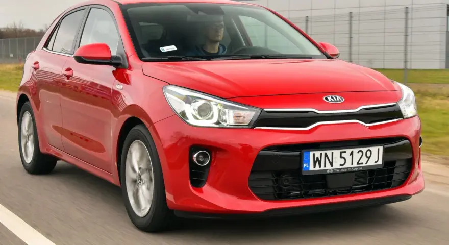 Czerwony Kia Rio w ruchu. Kierowca w okularach za kierownicą. Wiele pozytywnych kia rio opinie o tym modelu.