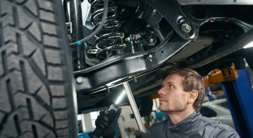 Mechanik bada elementy zawieszenia samochodu, oświetlając je latarką. Widoczna sprężyna i amortyzator.