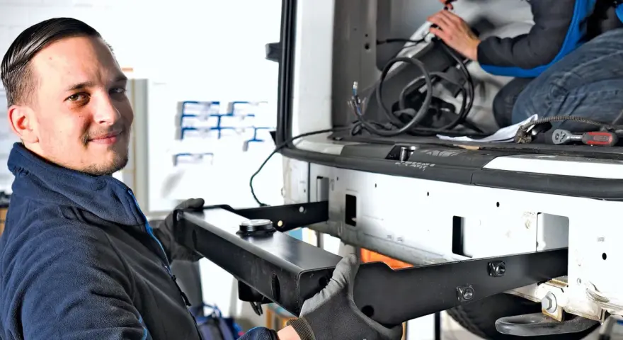 Mechanik z uśmiechem montuje hak w aucie. W tle widać drugą osobę pracującą przy instalacji.
