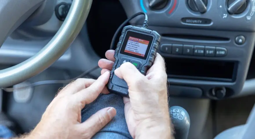 Ręce trzymające skaner diagnostyczny, który pokazuje menu z opcją "02 Sensor Test". To narzędzie pomoże zdiagnozować problemy z czujnikiem spalania stukowego.