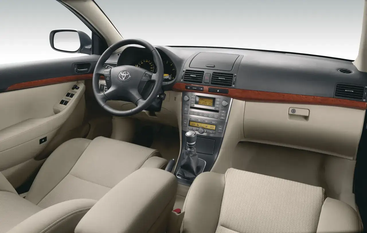 Wnętrze Toyoty Avensis T25: kierownica, deska rozdzielcza z radiem i klimatyzacją, dźwignia zmiany biegów, jasne fotele.