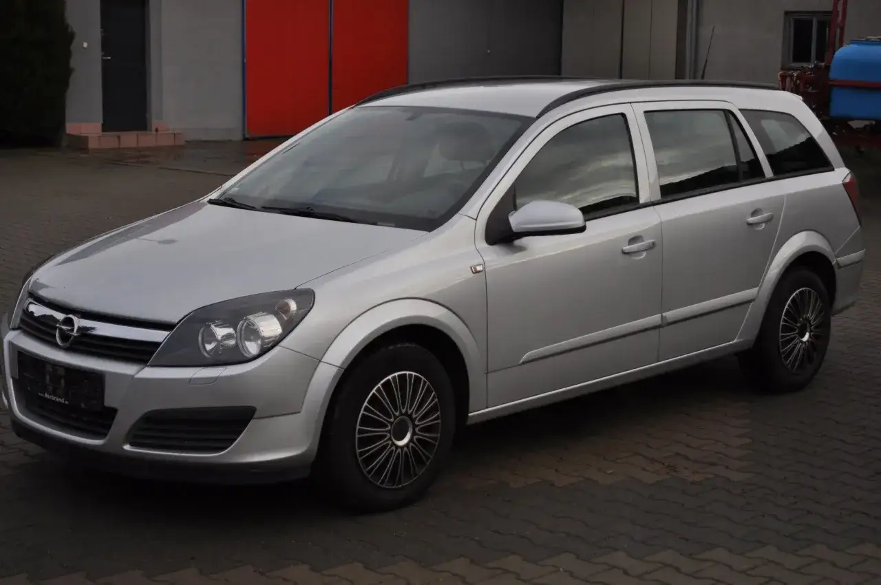 Srebrny Opel Astra H 1.6 benzyna kombi stoi na kostce brukowej przed budynkiem z czerwonymi drzwiami.
