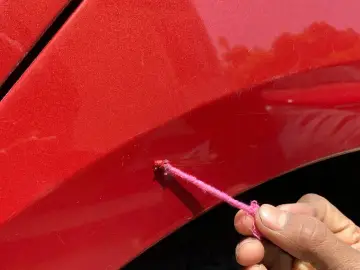 Ręka z różową nitką aplikuje zaprawki lakiernicze na rdzę na czerwonym błotniku samochodu.