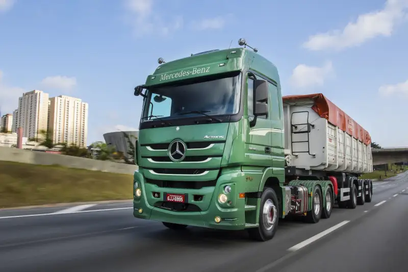 Zielony Mercedes Actros MP3 z naczepą na autostradzie, w tle budynki mieszkalne i most.
